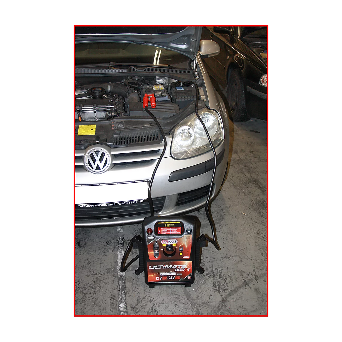 Ein silberner VW Golf mit geöffneter Motorhaube wird mit einem Starthilfegerät verbunden. Das Gerät steht vor dem Auto.